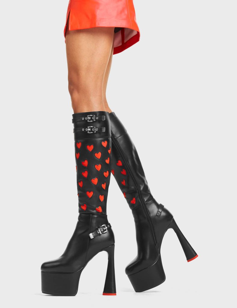 Talk Platform Knee High Boots in Black Stretch faux leather. These platform boots feature a panel of red heart-patterned material, with adjustable straps at the top and on the ankle of the boot. The straps have silver hardware including square-shaped buckles, and the platform sole features a red heart-shaped heel. Made with eco-friendly materials and 100% cruelty-free!